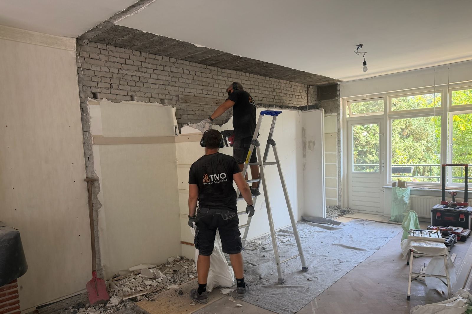 Twee vaklieden openen een draagmuur; de stalen ligger en de stempels staan al op hun plek