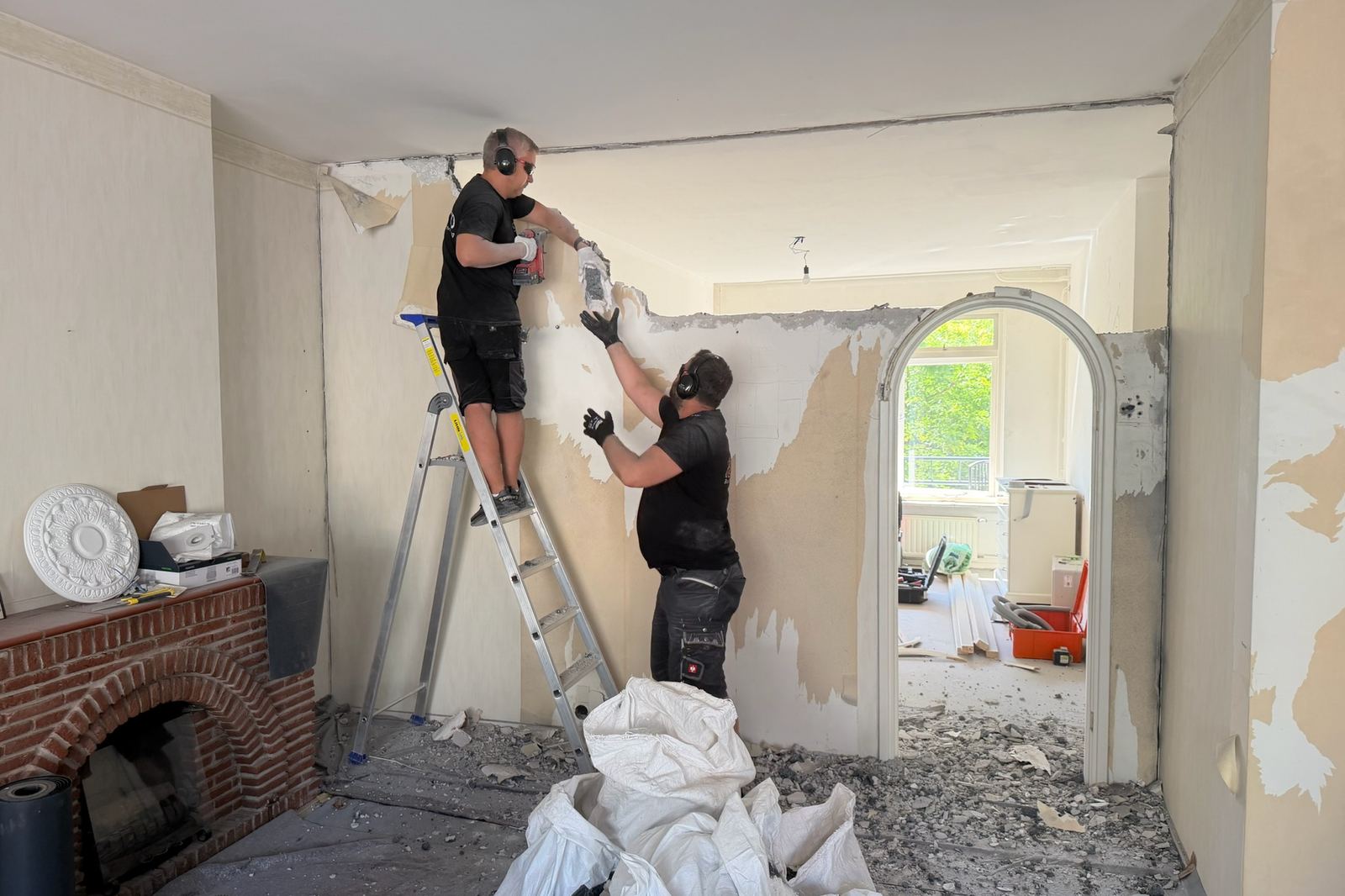 Vakmensen strippen een wand in een oudere woning met een gemetselde schouw en een boogdoorgang